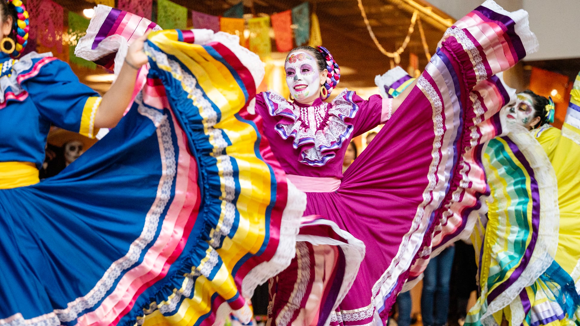 he dance group Mexicas performs in colorful traditional costumes with wide skirts spread out in motion. Several dancers wear face paint inspired by Día de los Muertos, and colorful paper decorations are visible in the background. The image depicts a vibrant scene focused on dance, culture, and movement.